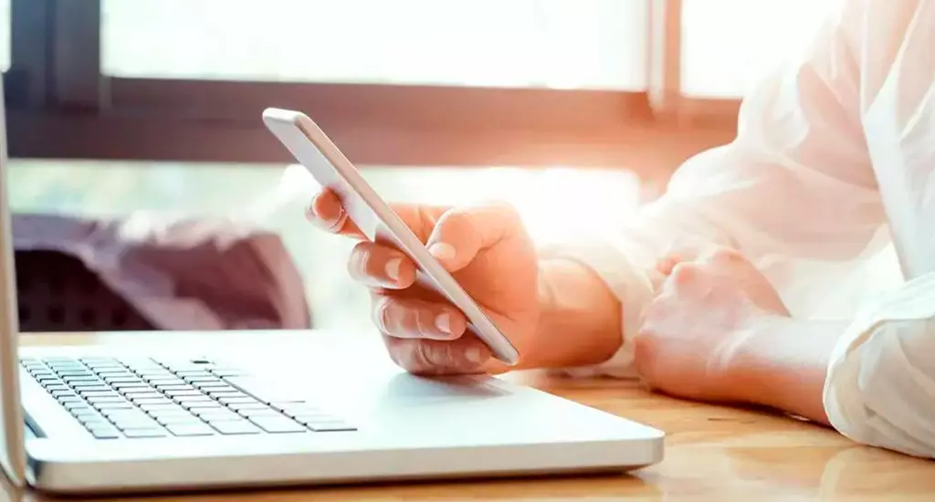 Pessoa utilizando smartphone enquanto trabalha em um laptop, com iluminação natural ao fundo. Ideal para estratégias de negócios, marketing digital e produtividade.
