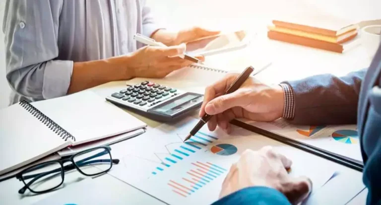 Duas pessoas sentadas à mesa analisando relatórios financeiros com gráficos de barras e pizza; uma delas aponta para os dados com uma caneta enquanto há uma calculadora, caderno e óculos sobre a mesa.