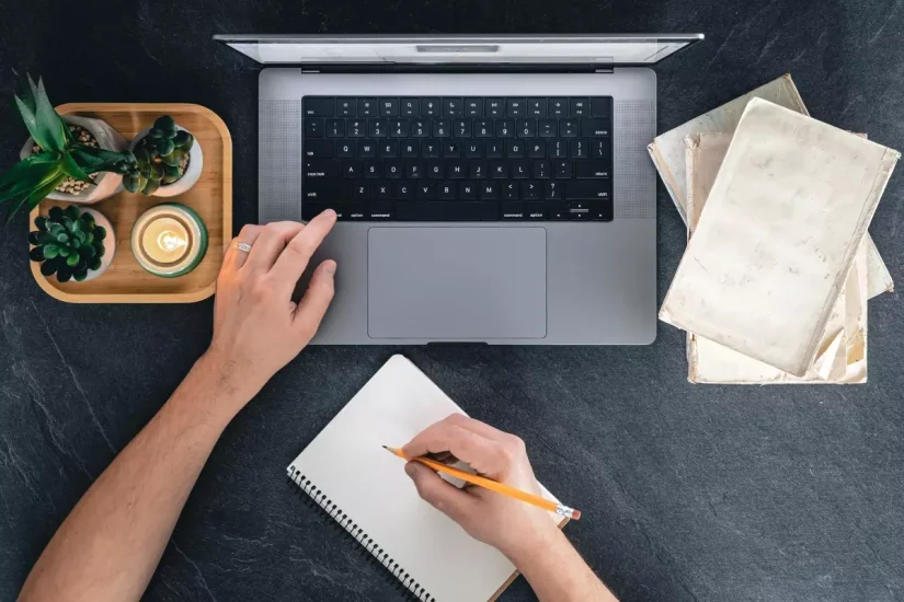 Imagem de pessoa escrevendo em caderno com laptop aberto, plantas e livros ao redor, ideal para aprender como escrever conteúdo para SEO de forma eficiente.