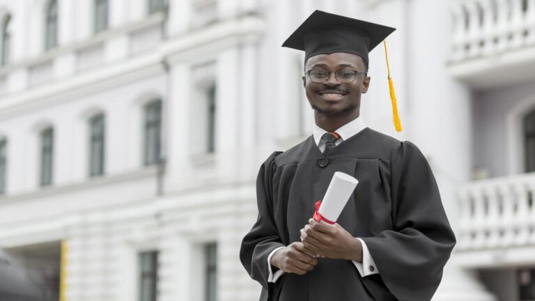 Um jovem negro sorridente vestido com beca e capelo de formatura preto segura seu diploma enrolado com um laço vermelho.
