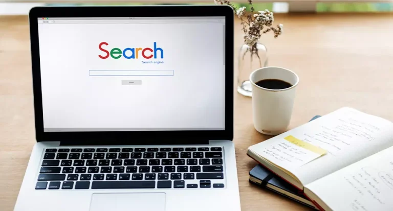 Imagem de um laptop aberto exibindo a página do Google Search, ao lado de uma xícara de café, um caderno com anotações e plantas decorativas, ideal para pesquisas na internet.