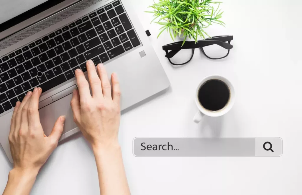 Pessoa pesquisando 'o que é SERP' em um laptop, ao lado de uma xícara de café, óculos e uma planta de cabelo verde. Imagem ilustrativa para SEO.