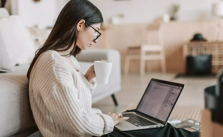 Mulher jovem trabalhando no laptop e segurando uma caneca em sala de estar moderna.