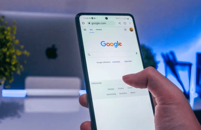 Mão segurando um smartphone com a página inicial do Google aberta no navegador, com um laptop desfocado ao fundo sobre a mesa.
