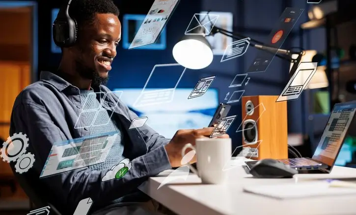 Homem negro sorridente usando fones de ouvido e trabalhando em um tablet em uma mesa com um notebook. Várias telas e ícones digitais flutuam ao redor dele.