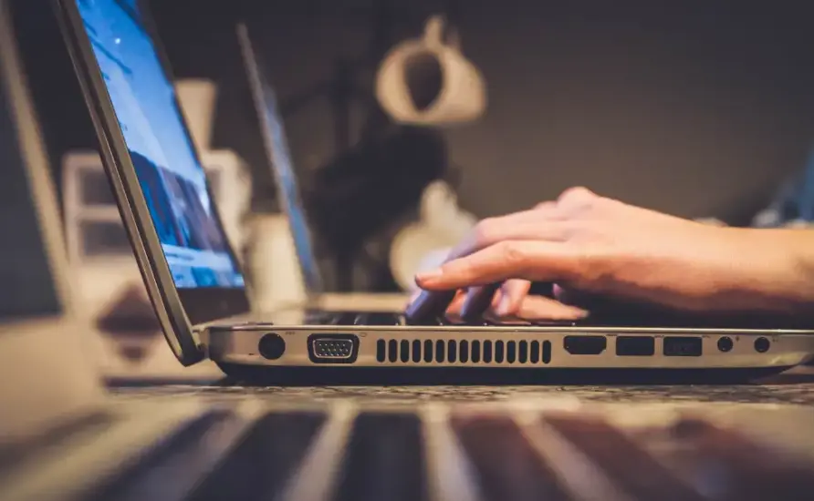 Pessoa digitando em um notebook aberto sobre uma mesa, com foco nas mãos e no teclado, em um ambiente interno e iluminado.