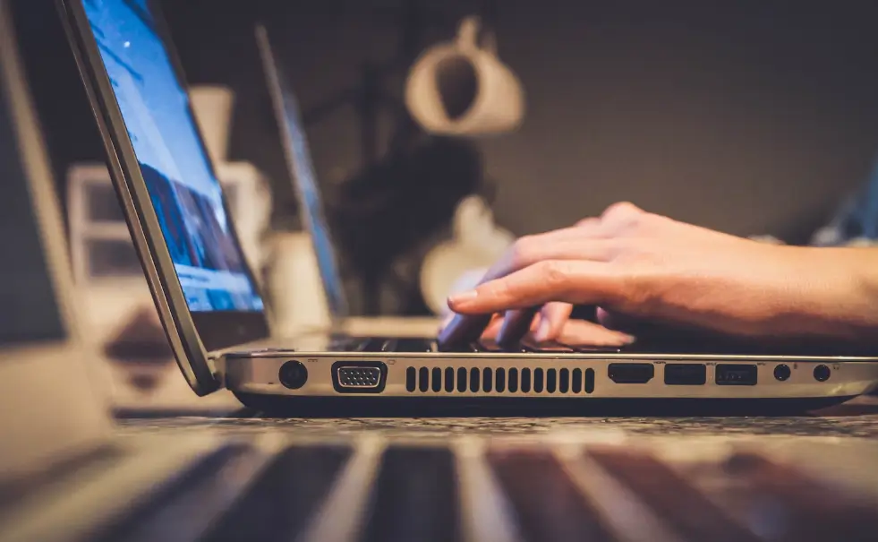 Pessoa digitando em um notebook aberto sobre uma mesa, com foco nas mãos e no teclado, em um ambiente interno e iluminado.