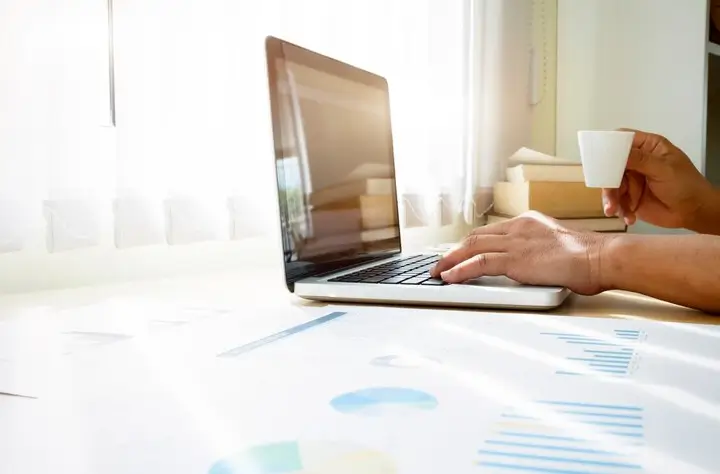 Pessoa trabalhando em um laptop com uma xícara de café, com gráficos e relatórios na mesa. A cena simboliza o trabalho analítico necessário para entender Como fazer pesquisa de palavras-chave para SEO