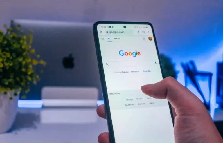 Smartphone exibindo a página inicial do Google em um ambiente com iluminação azul. Uma mão segura o aparelho e um dedo toca a tela, sugerindo uma interação com a pesquisa ou navegação.
