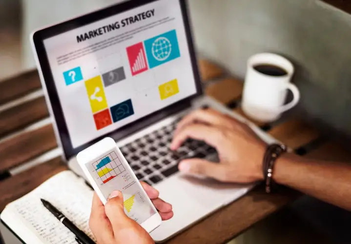 Pessoa utilizando smartphone e laptop em uma mesa de trabalho, com gráfico e estratégia de marketing na tela, analisando se vale a pena investir em SEO.