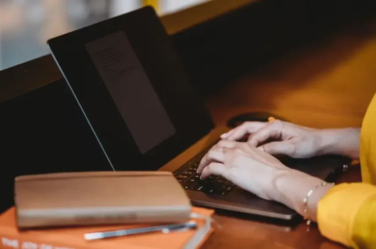 Uma vista aproximada das mãos de uma pessoa digitando no teclado de um laptop. A pessoa está vestindo uma camisa amarela e uma pulseira. Na mesa, há um caderno marrom, uma caneta e uma pilha de livros com capas laranjas. A iluminação é quente e o fundo está desfocado.