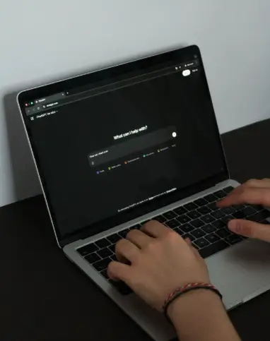Mãos digitando em um laptop, mostrando a tela do ChatGPT com a pergunta "What can I help with?" (Com o que posso ajudar?), ilustrando o uso de inteligência artificial.