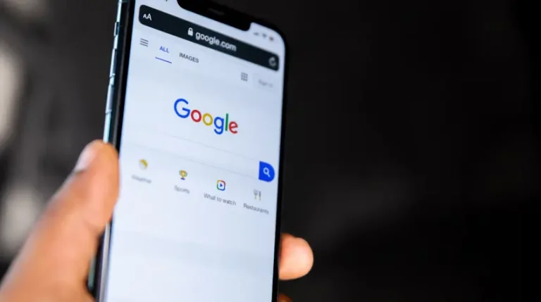 Barra de pesquisa e o logotipo do Google, com cores vivas, são o foco principal. O plano de fundo está desfocado em tons escuros, direcionando a atenção para o aparelho e a busca online.