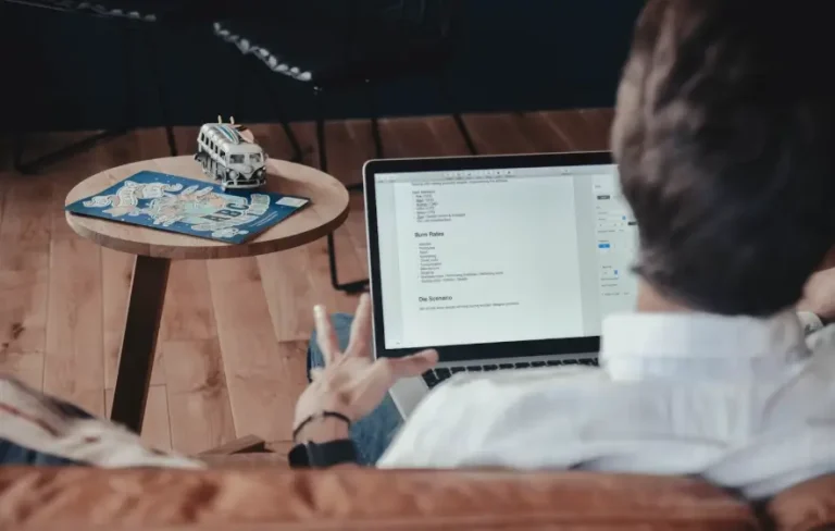 Vista das costas de um homem sentado em um sofá marrom, olhando para a tela de um laptop. O homem está vestindo uma camisa branca. No chão de madeira, ao lado dele, há uma pequena mesa redonda de madeira com uma miniatura de kombi e um mapa azul. A tela do laptop mostra um documento com texto.