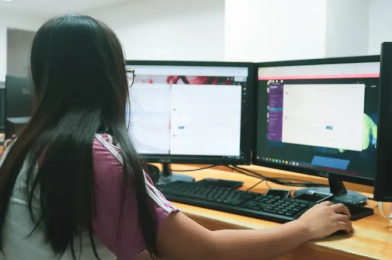Mulher de costas usando dois monitores de computador para analisar dados e códigos. A imagem simboliza o trabalho técnico e analítico de Otimização SEO para imagens em blogs e de websites.