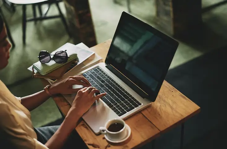 Uma pessoa sentada em uma mesa, digitando em um notebook. Ao lado do laptop, há uma xícara de café, uma pilha de cadernos. A imagem é vista por cima do ombro da pessoa, focando nas mãos digitando no teclado.