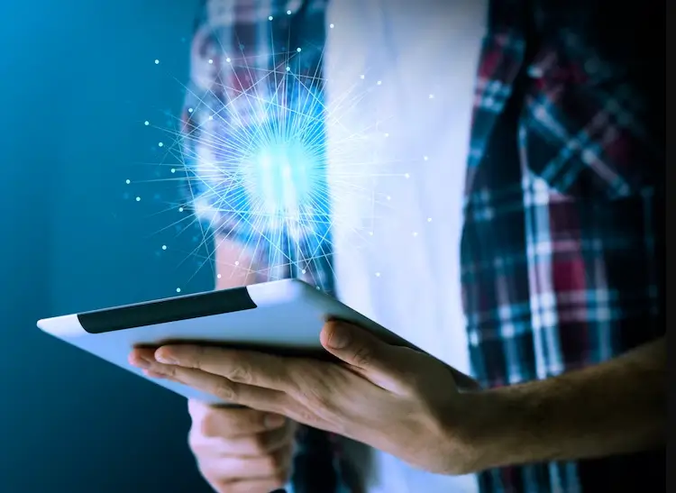 Homem de camisa xadrez e camiseta branca segurando um tablet. Uma luz azul brilhante, com um efeito de explosão e partículas, se projeta da tela do tablet em direção às suas mãos e ao seu corpo. A imagem simboliza tecnologia, inovação e a transferência de dados ou ideias.