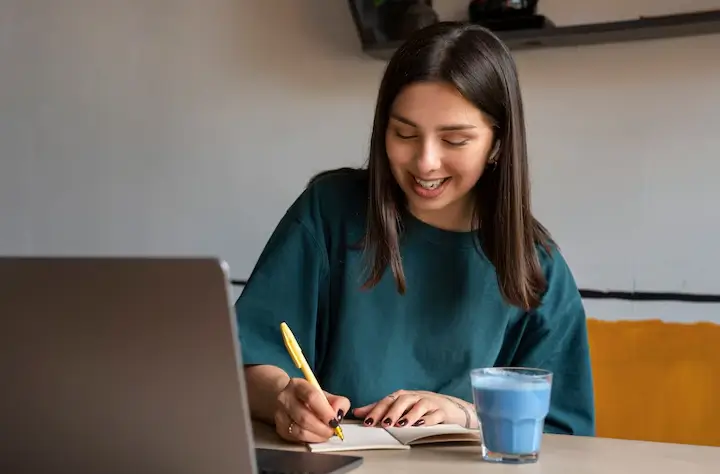 Mulher jovem estudando ou trabalhando em casa com um laptop, anotando em um caderno informacoes sobre como se manter atualizado em SEO em 2025 com uma caneta amarela, ao lado um copo de bebida azul, sorrindo e focada na tarefa.
