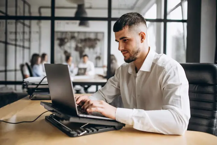 Homem está em um ambiente de escritório com uma sala de reunião de vidro ao fundo, onde outras pessoas estão sentadas. A imagem transmite uma sensação de trabalho focado e profissionalismo.