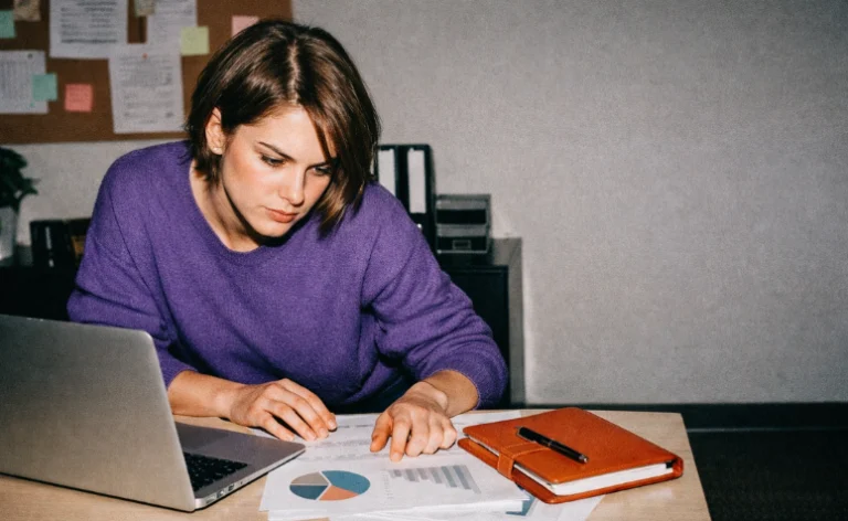 Mulher de blusa roxa analisando relatórios com gráficos sobre a mesa de escritório. Ao lado, há um notebook aberto e uma agenda marrom com caneta.