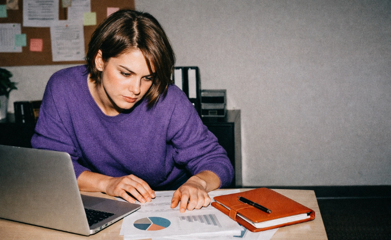Mulher de blusa roxa analisando relatórios com gráficos sobre a mesa de escritório. Ao lado, há um notebook aberto e uma agenda marrom com caneta.